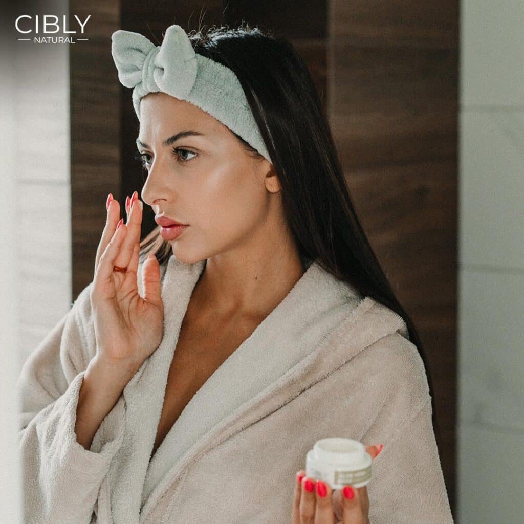 Жена нанася крем против зачервяване и раздразнена кожа, носейки пухкав халат и лента за коса, с продукт от Cibly.