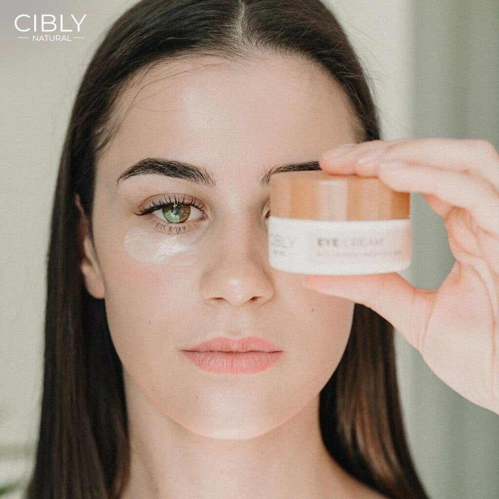 Жена демонстрира как се нанася околоочен крем, държейки продукт на Cibly и с нанесен крем под окото.